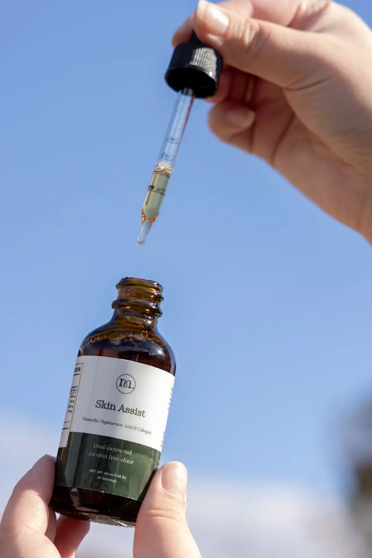 Hand holding a dropper over a bottle labeled 'Skin Assistor' against a clear blue sky.