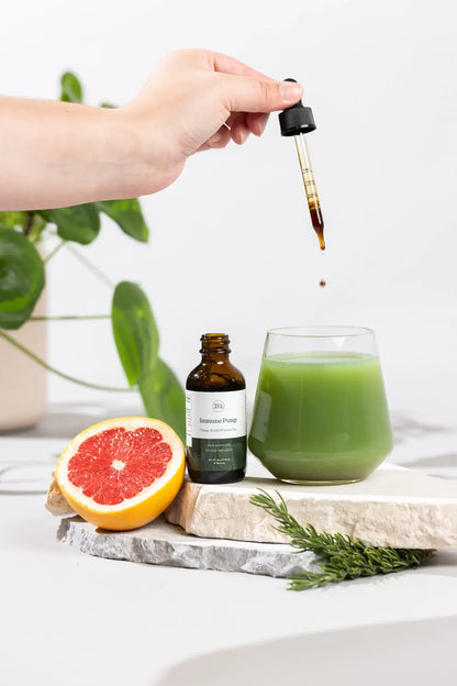 Hand pouring liquid from a dropper into a glass of green juice with a bottle and grapefruit on a white surface.