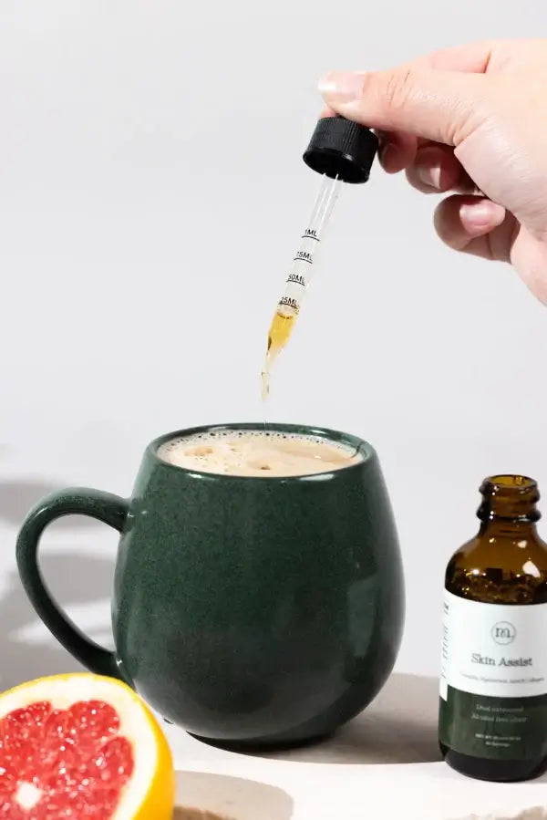 Hand dropping a liquid into a mug with a bottle labeled 'Skin Assistant' on a white background