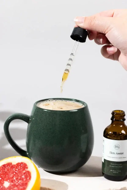 Hand dropping a liquid into a mug with a bottle labeled 'Skin Assistant' on a white background