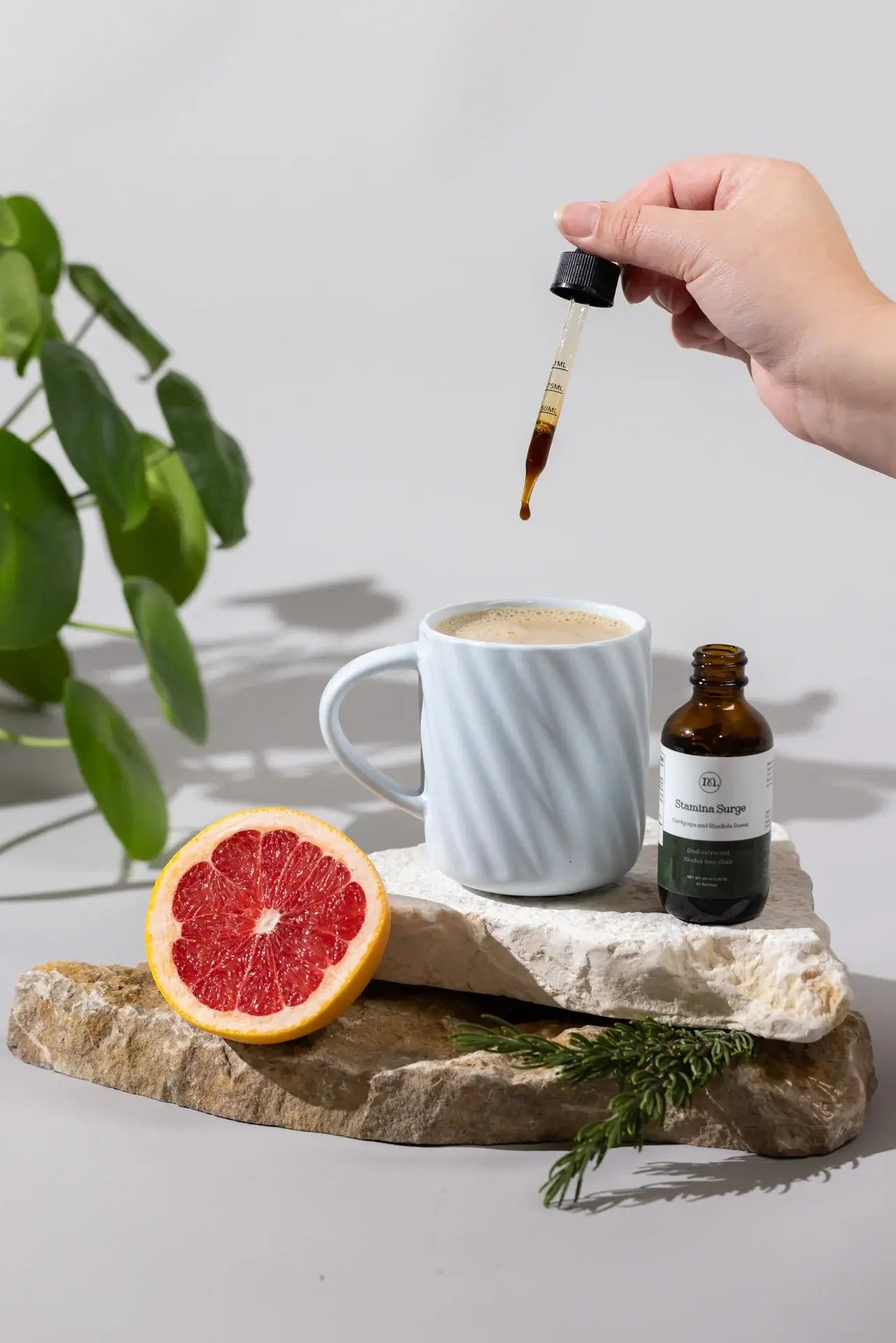 Hand holding a dropper over a cup of coffee with a bottle and grapefruit on a stone surface.