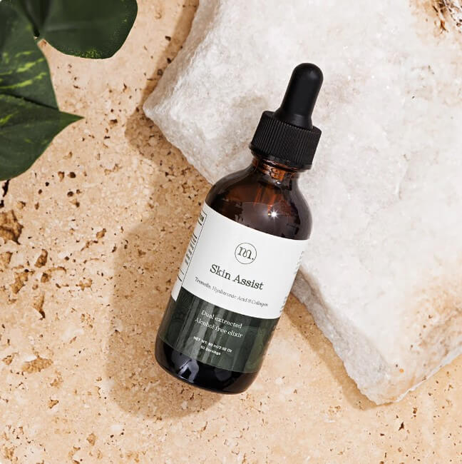 Skincare dropper bottle labeled 'Skin Assist' on a textured surface with a leaf in the corner.